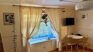 Une pièce avec une fenêtre, des rideaux, un téléviseur, une table avec des chaises et un ventilateur de plafond.