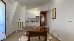 Un comedor con una mesa de madera y cuatro sillas, una cocina al fondo y una pared blanca con un cuadro enmarcado.