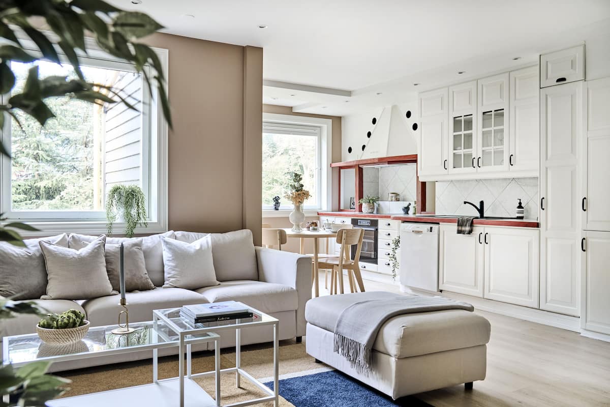 Un salon moderne avec un canapé, une table basse et un pouf. On y trouve également un coin cuisine avec des meubles blancs et une table à manger.