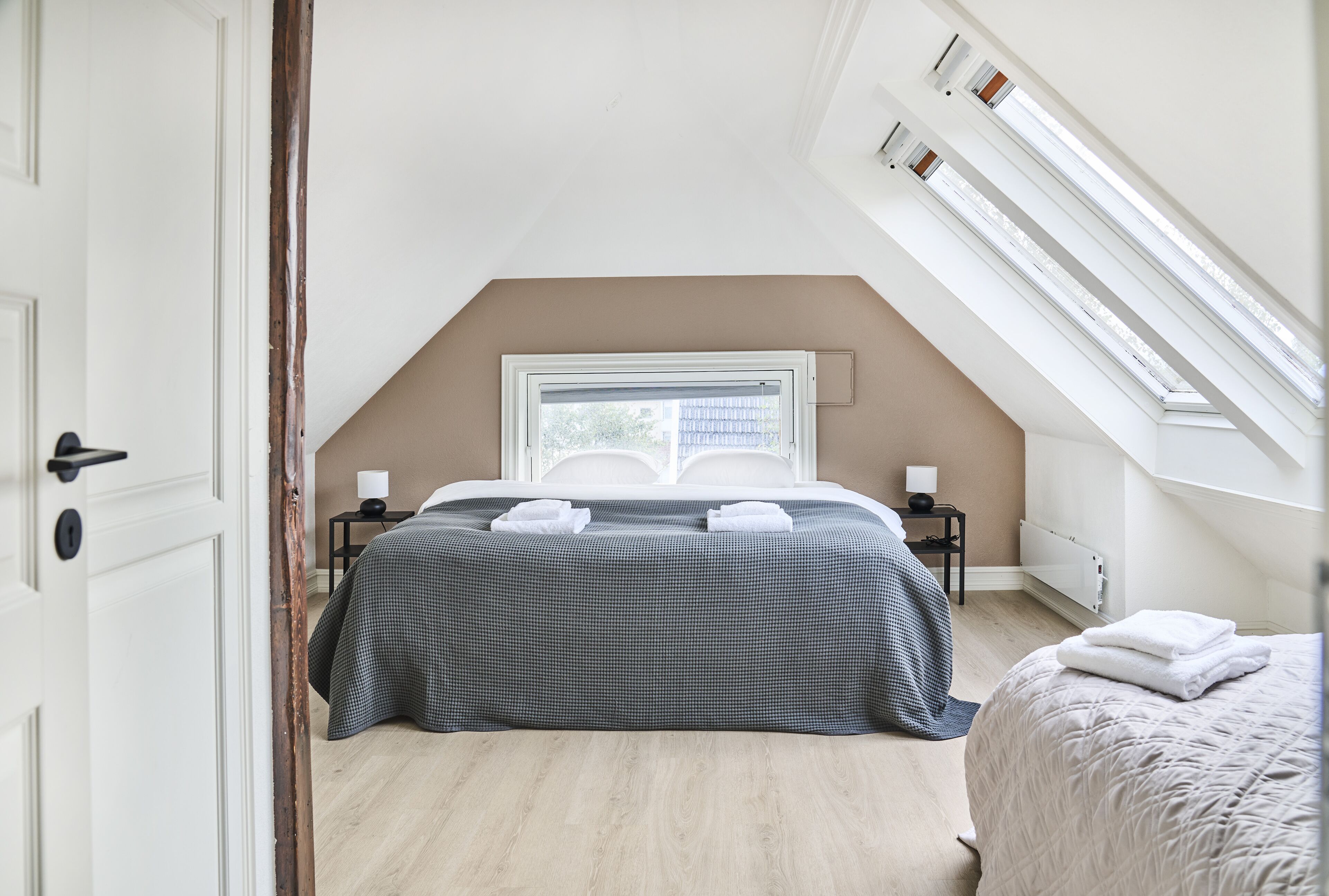 Un dormitorio con techo inclinado, una cama con colcha de cuadros, una mesita de noche con lámpara y una ventana con persianas.