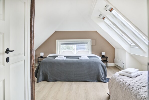 Un dormitorio con techo inclinado, una cama con colcha de cuadros, una mesita de noche con lámpara y una ventana con persianas.