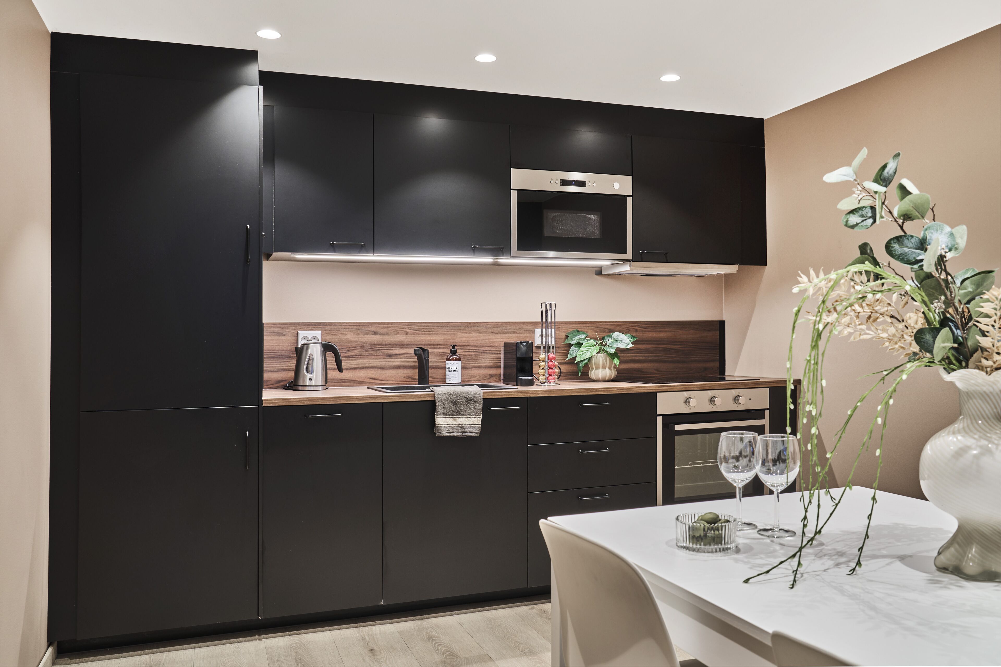 A modern kitchen with black cabinets, a wooden countertop, a built-in oven, and a dining area with a table and chairs.