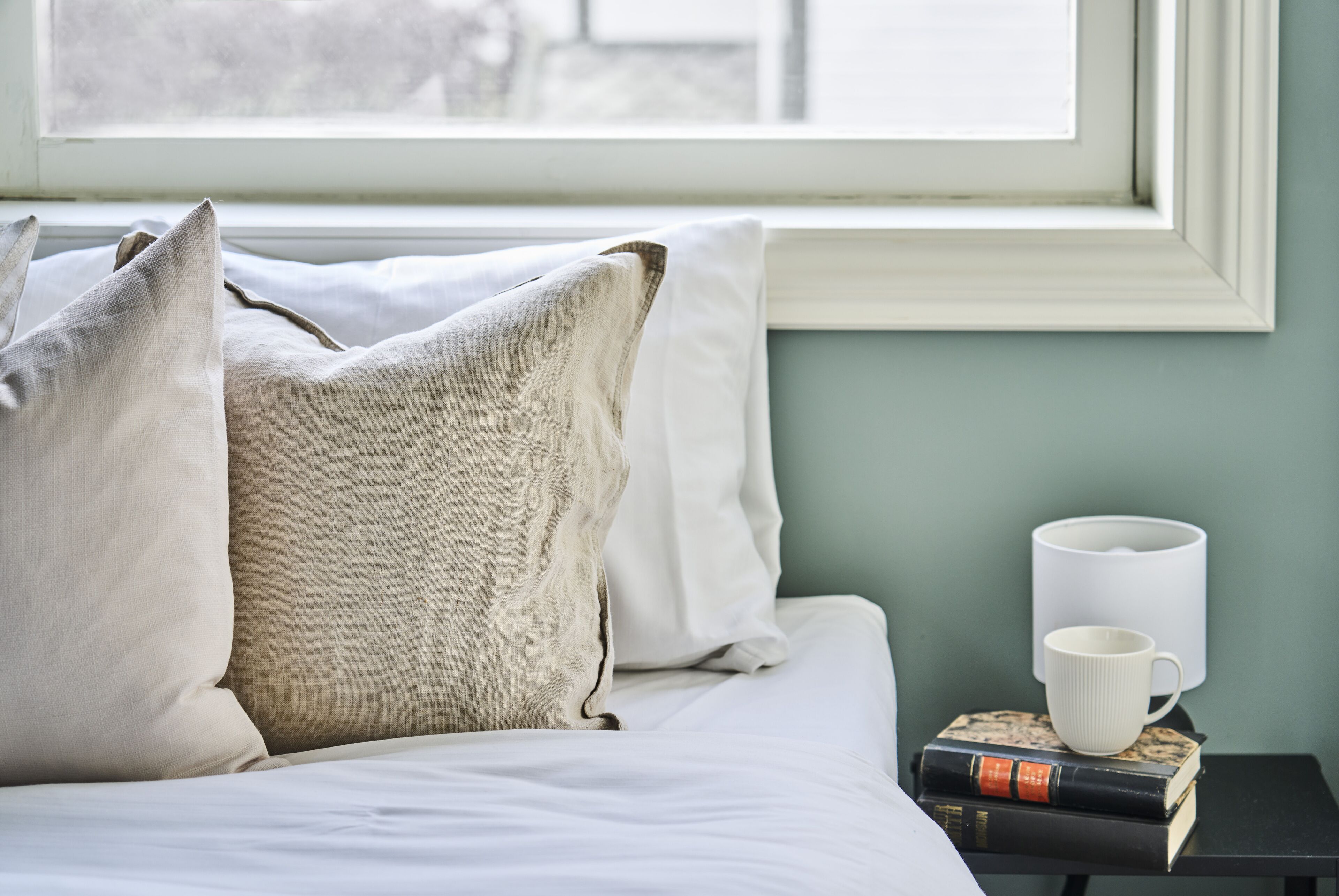A neatly made bed with pillows, a white mug, and books on a nightstand by a window.