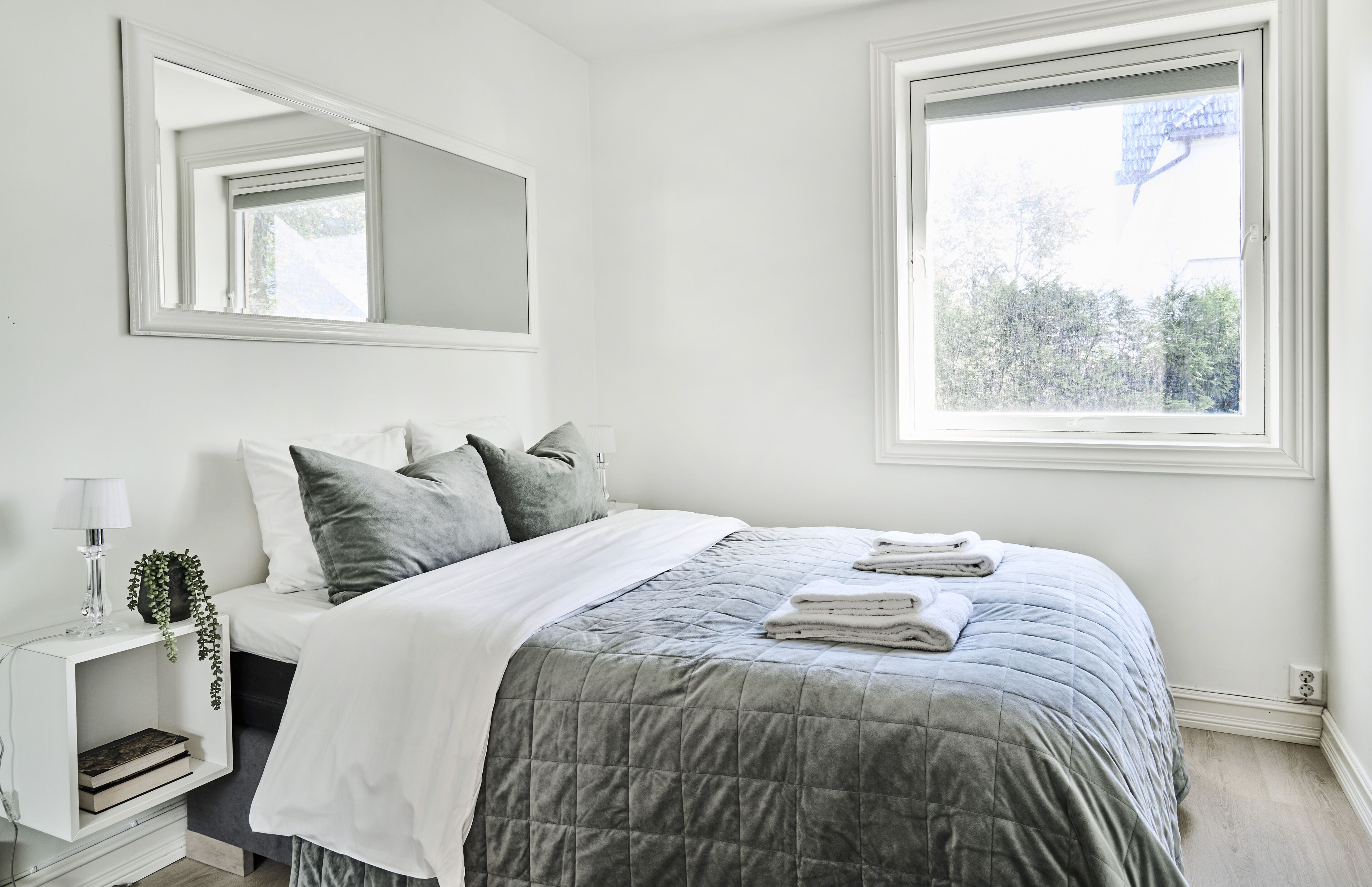 A bedroom with a bed, a bedside table with a lamp, a mirror, and a window.