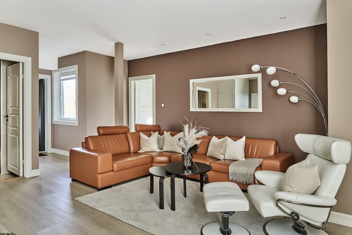 Un salon moderne avec un canapé d’angle en cuir marron, un fauteuil blanc, une petite table ronde et un grand miroir accroché au mur.