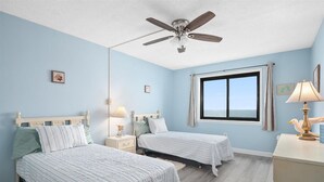 Un dormitorio con dos camas, ventilador de techo, ventana con vistas y escritorio con lámpara.