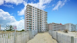Un hotel frente a la playa con un camino de arena que lleva a la entrada, un cartel que indica el paso por una duna y un cielo azul despejado.