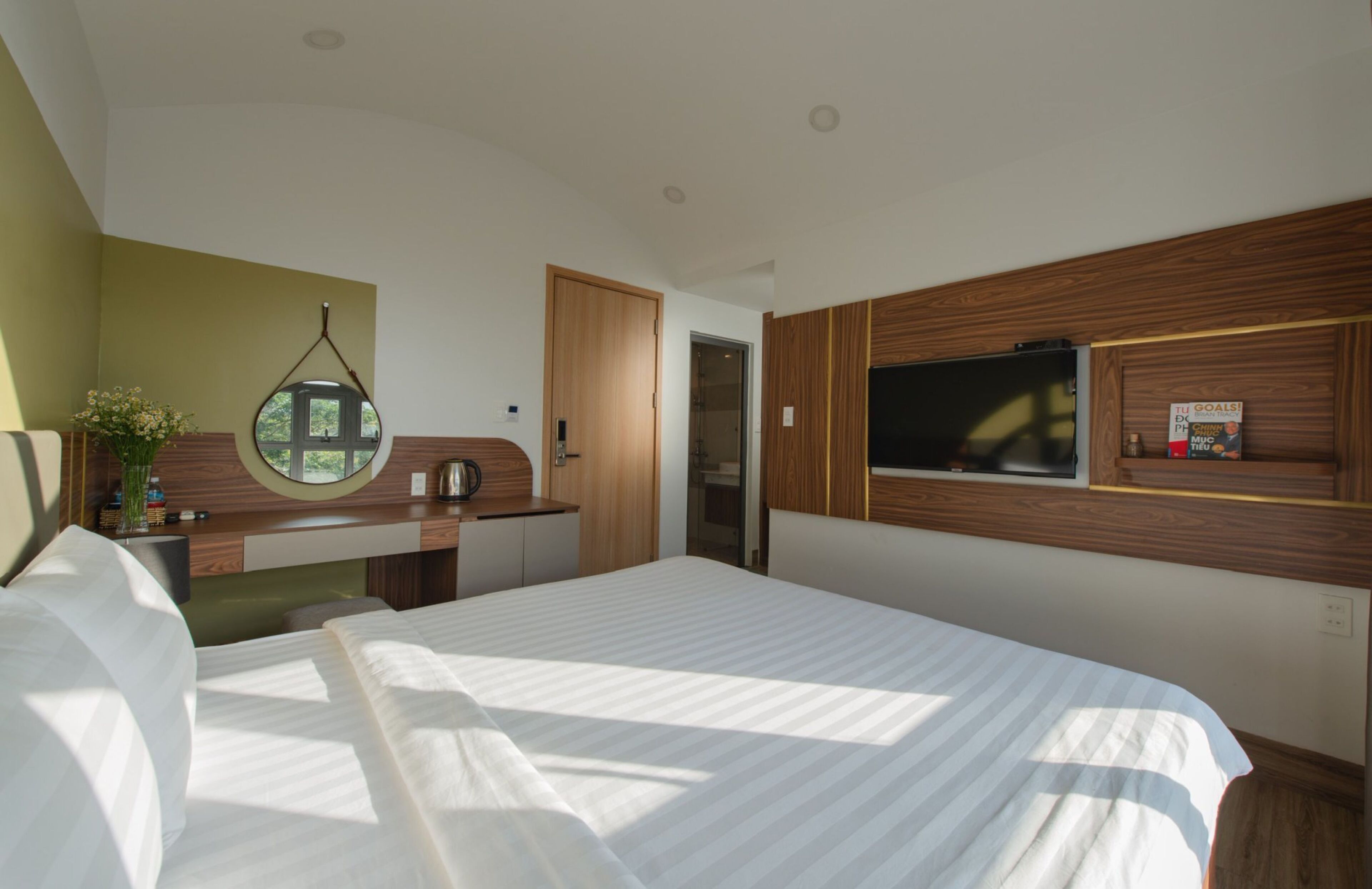 Un dormitorio moderno con una cama grande, cabecera de madera, un espejo redondo, un escritorio pequeño y un televisor montado en la pared.