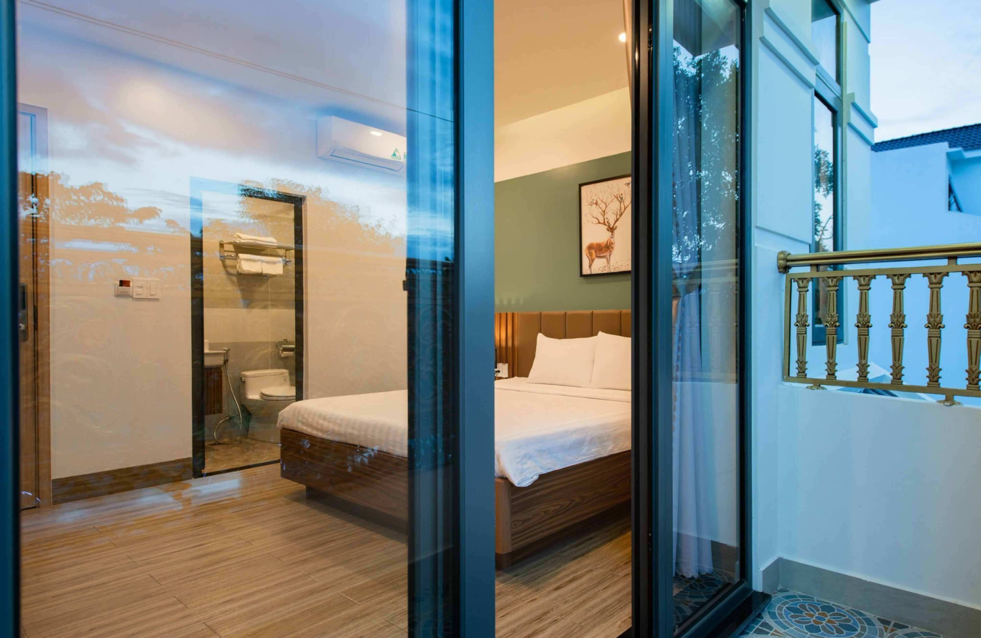 A modern hotel room with a glass partition, a wooden floor, a bed with white linens, a framed deer picture on the wall, and a balcony with a railing.