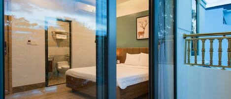 A modern hotel room with a glass partition, a wooden floor, a bed with white linens, a framed deer picture on the wall, and a balcony with a railing.