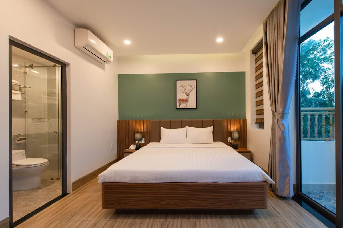 Quarto de hotel moderno com uma cama grande, chuveiro box de vidro, parede verde decorativa e vista para o exterior.