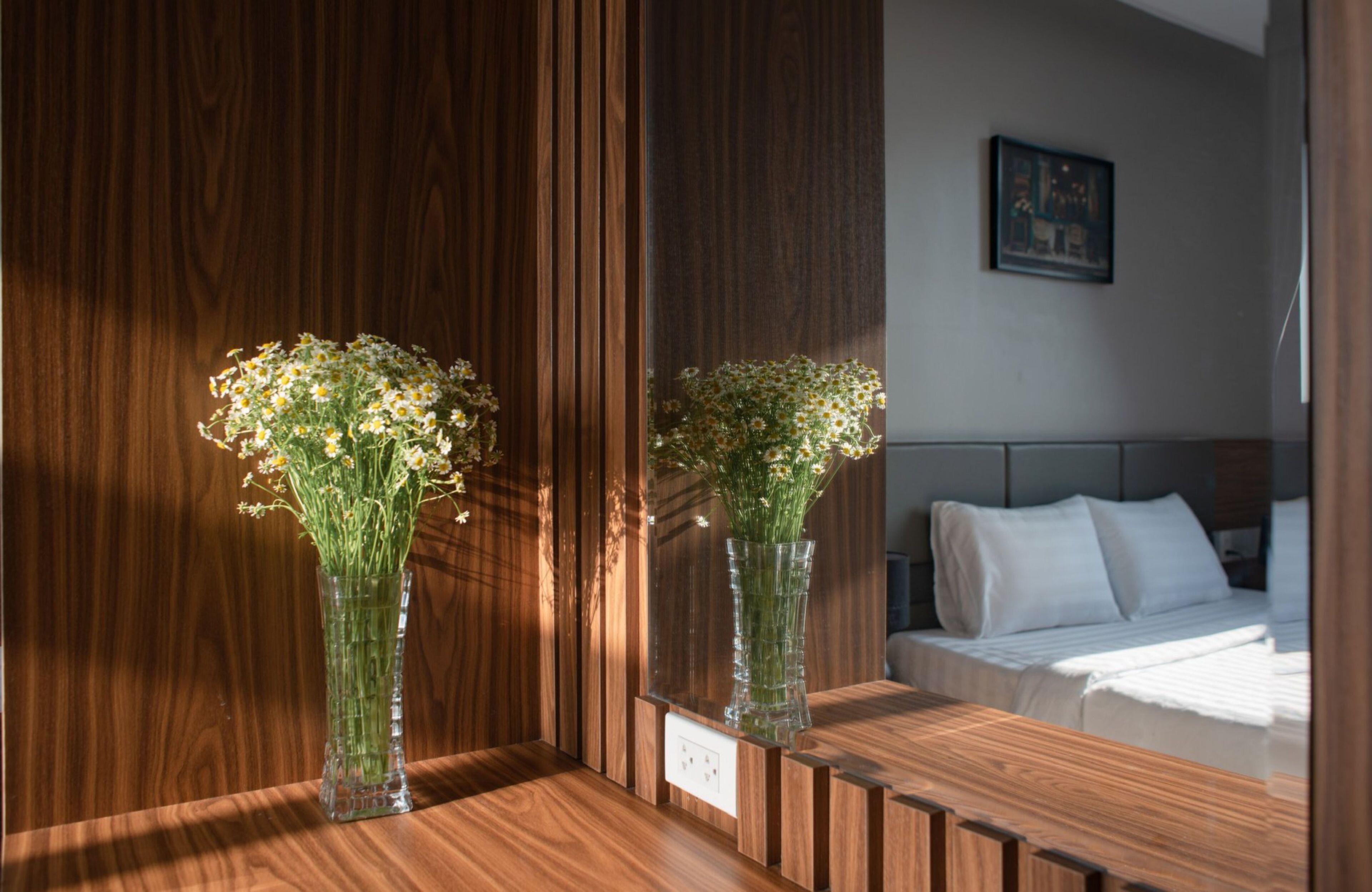 Une chambre d’hôtel avec un mur lambrissé en bois, un lit avec une literie blanche et deux vases transparents ornés de fleurs, posés sur une table de chevet en bois.