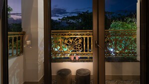 Un balcón con barandillas de metal ornamentadas, dos taburetes redondos y vistas a árboles y al atardecer.