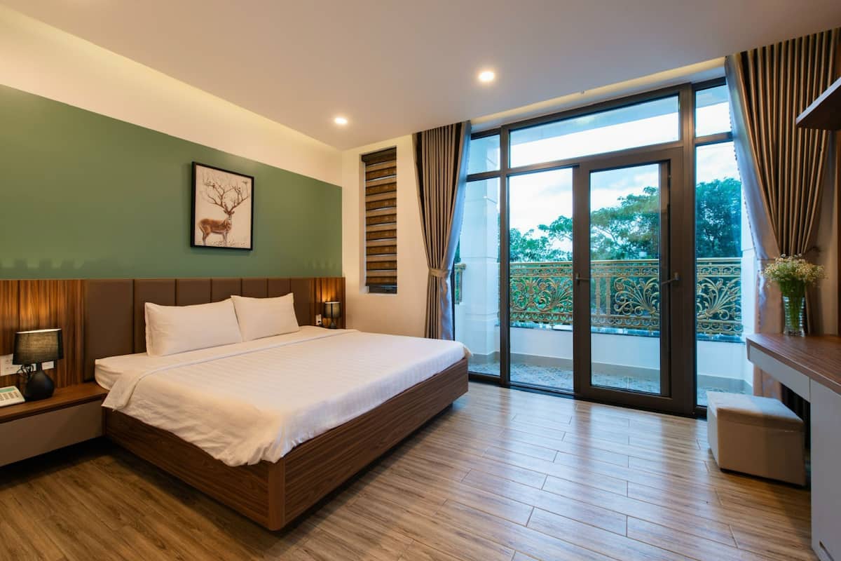 Quarto de hotel moderno com uma cama grande, uma mesa de cabeceira com abajur, um quadro de um veado na parede e uma varanda com vista para árvores.