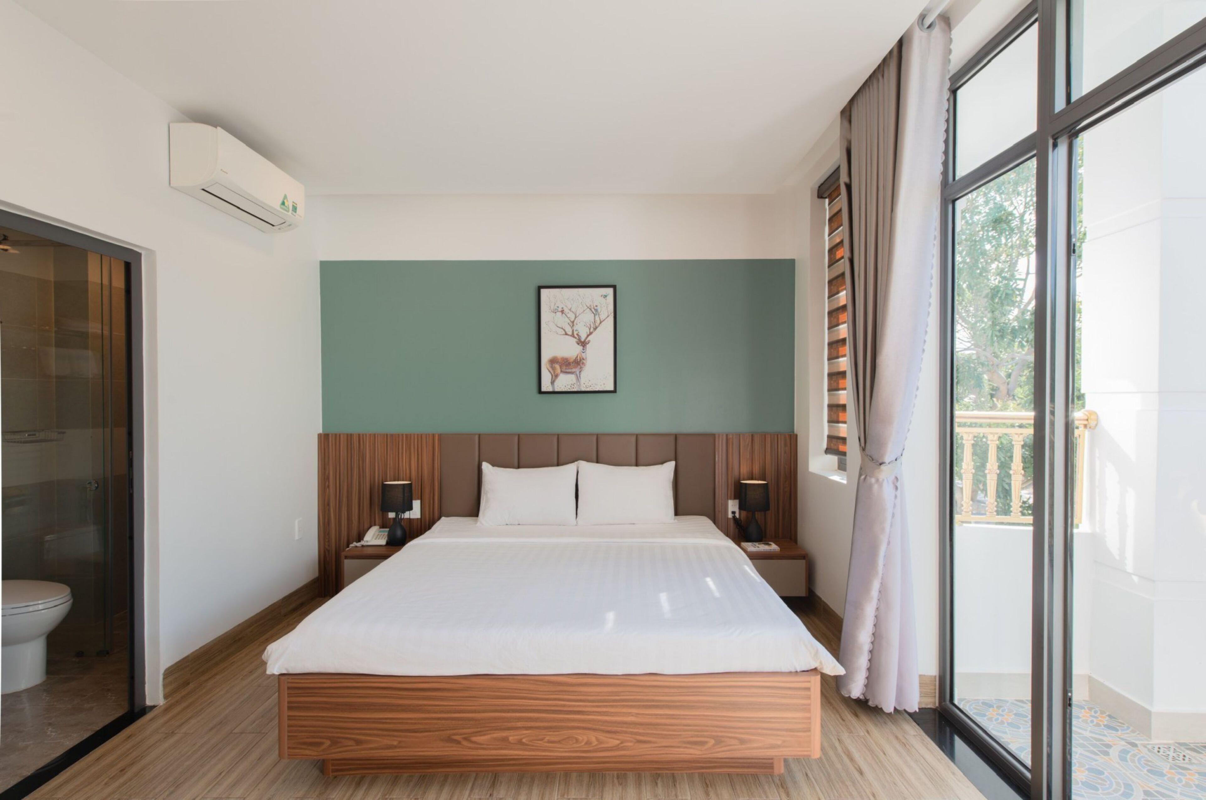 A modern hotel room with a large bed, wooden headboard, a framed picture on the wall, and a balcony with curtains.