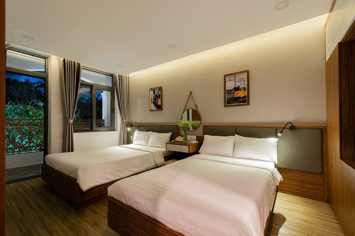 Quarto de hotel com duas camas, uma janela grande com cortinas, uma mesa de cabeceira com abajur e obras de arte na parede.