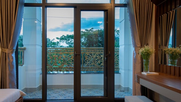 Una habitación con puerta de vidrio corredera que da a un balcón con barandilla decorativa. Hay una cama y un pequeño banco en la habitación.
