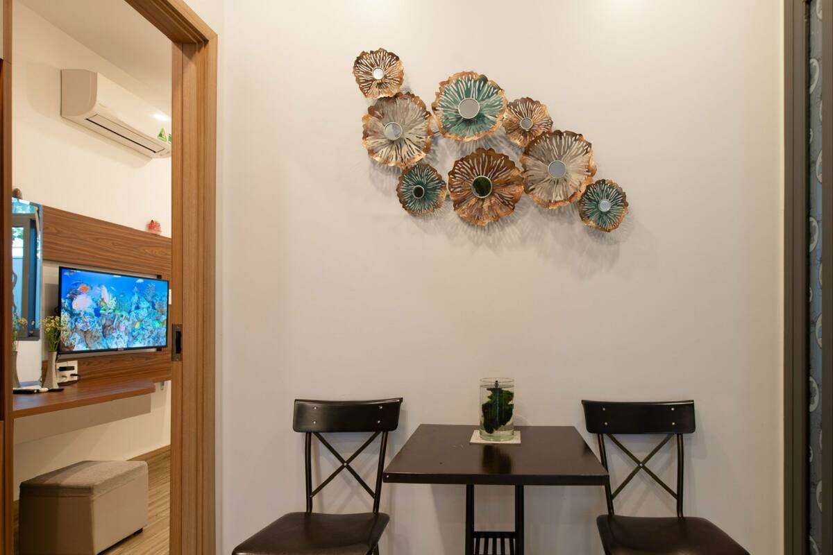 Quarto com televisão em prateleira de madeira, duas cadeiras, uma mesa e arte decorativa na parede.