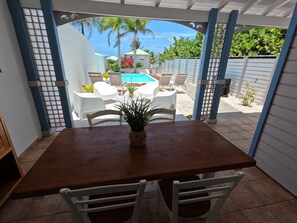 Une terrasse avec une table en bois et des chaises blanches, donnant sur un espace piscine avec des chaises longues et une petite maison en arrière-plan.