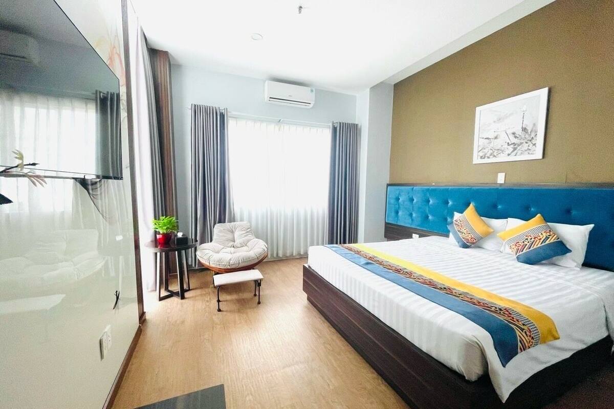 Een hotelkamer met een groot bed, een stoel, een tafeltje met een plant en een raam met gordijnen.
