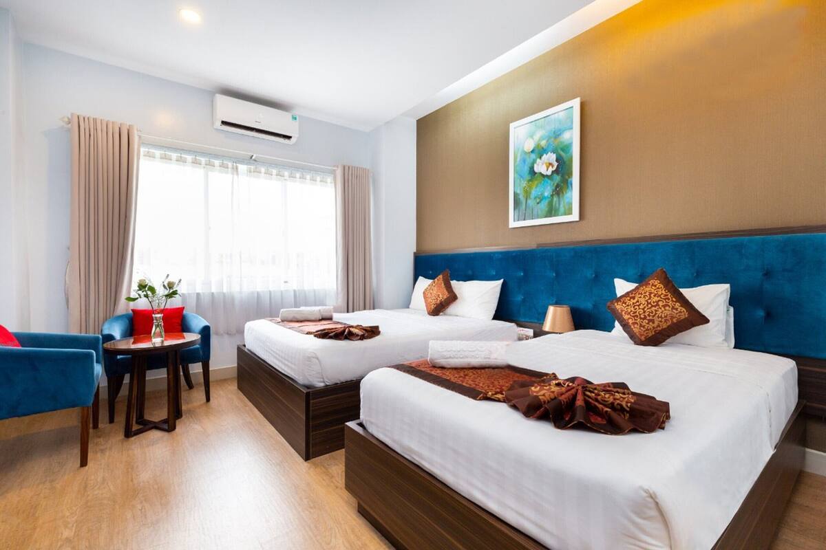 Een hotelkamer met twee bedden, een blauw bekleed hoofdeinde, een schilderij aan de muur en een raam met gordijnen.
