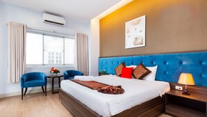 Une chambre d’hôtel avec un grand lit, une tête de lit bleue, une table de chevet avec une lampe, une petite table ronde ornée d’un vase de fleurs et une fenêtre avec des rideaux.