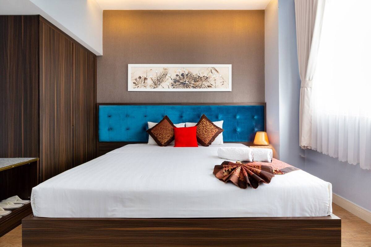 Een groot bed met een blauw hoofdeinde, witte beddengoed en meerdere decoratieve kussens. Daarboven hangt een kunstwerk in een lijst en er staat een lamp op het nachtkastje.