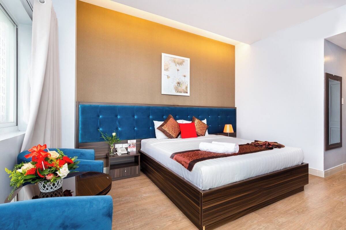 Een moderne hotelkamer met een groot bed, een blauw beklede hoofdeinde, een nachtkastje met een lamp en een raam met gordijnen.