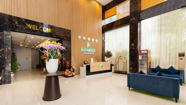 Un hall d’hôtel avec un comptoir d’accueil, une pancarte « BIENVENUE », une pancarte « Bamboo », un canapé bleu et une plante en pot.