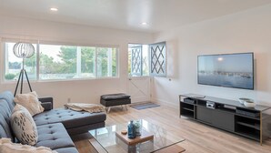 A modern living room with a grey sectional sofa, a glass coffee table, a black ottoman, a TV on a black stand, and large windows with a view of greenery. - 3896 Gather and Rest - Mountain Views, Large Patio (Del Rey Oaks)