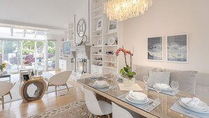 Ein moderner Essbereich mit einem Glastisch, weißen Stühlen und einer Chandelier-Leuchte. An den Wänden befinden sich Regale mit Dekoration und gerahmte Bilder.