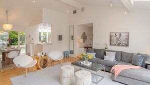 Ein modernes Wohnzimmer mit einem grauen Sofa, weißen Stühlen und einem Couchtisch. Es gibt einen Essbereich mit einem weißen Tisch und Stühlen. Der Raum verfügt über große Fenster und eine Chandelier.