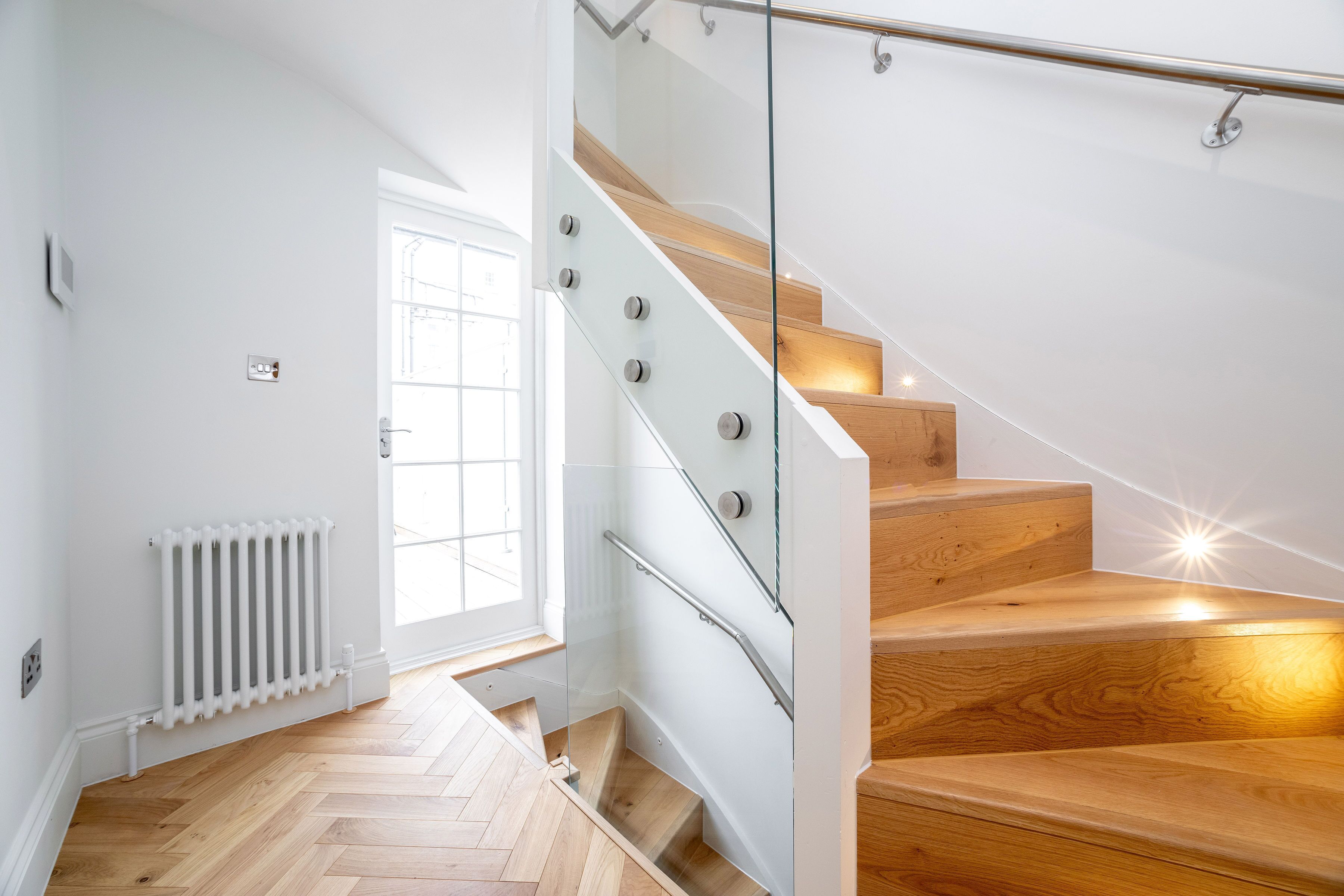 A modern staircase with glass railings and wooden steps, leading to a bright room with a white radiator and a window.