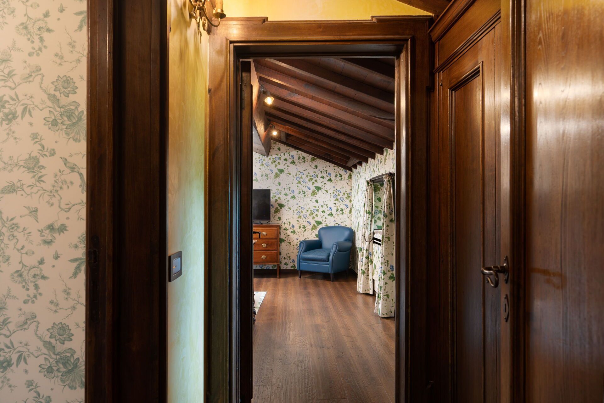 Un pasillo estrecho con puertas y paredes de madera, que conduce a una habitación con papel tapiz floral y un sillón azul.