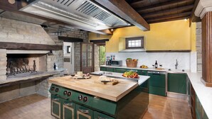 A kitchen with a central island, a stone fireplace, and wooden beams on the ceiling. - Villa Torre delle Rose by Geco Vacation Rentals (Visignano)