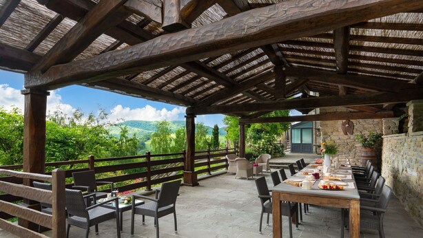 An outdoor patio with stone flooring, wooden furniture, and a view of a lush landscape. - Villa Torre delle Rose by Geco Vacation Rentals (Visignano)