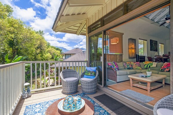 A spacious balcony with wicker furniture, a glass door leading to a living area with a sofa and coffee table, and a view of greenery and mountains.