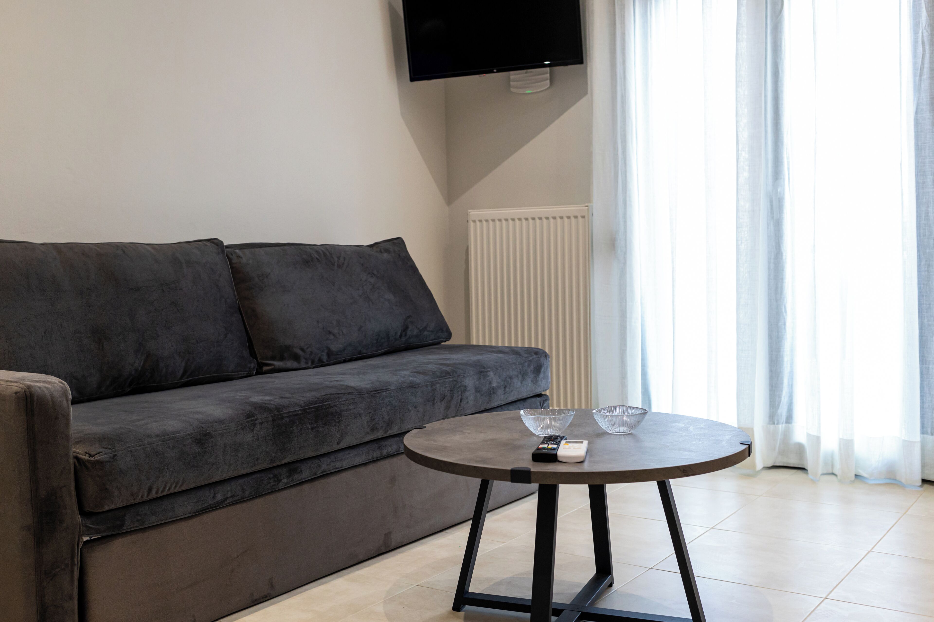 A modern living room with a grey sofa, a round table with two glass bowls, a remote control, and a television mounted on the wall.