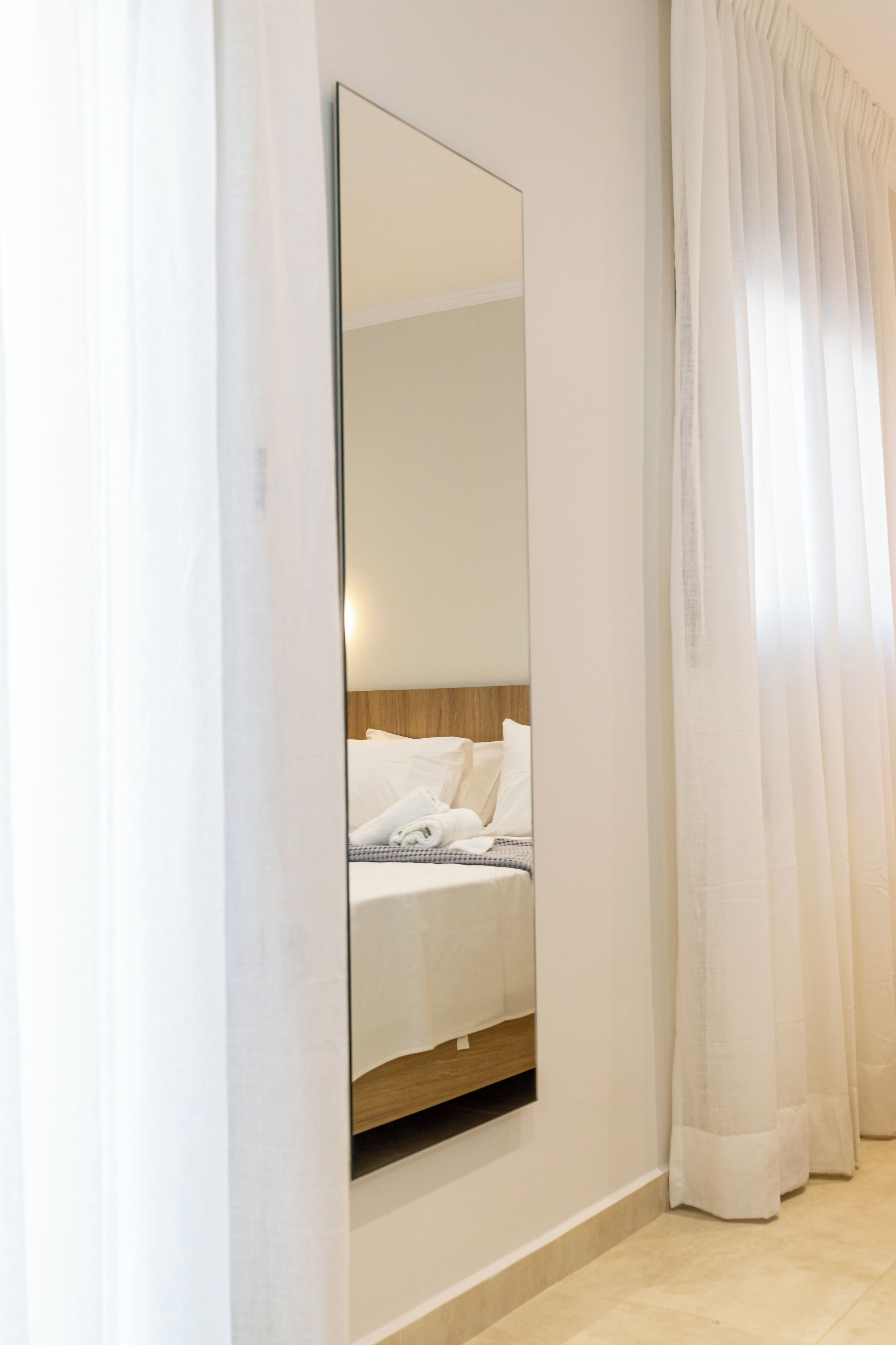 A hotel room with a large mirror, a bed with white linens, and sheer curtains.