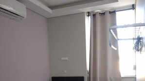 A small, clean room with a bed, a desk, and a chair. There is a window with curtains, a small table, and a white cabinet. - Ipê Rosa Hotel (Bauru)