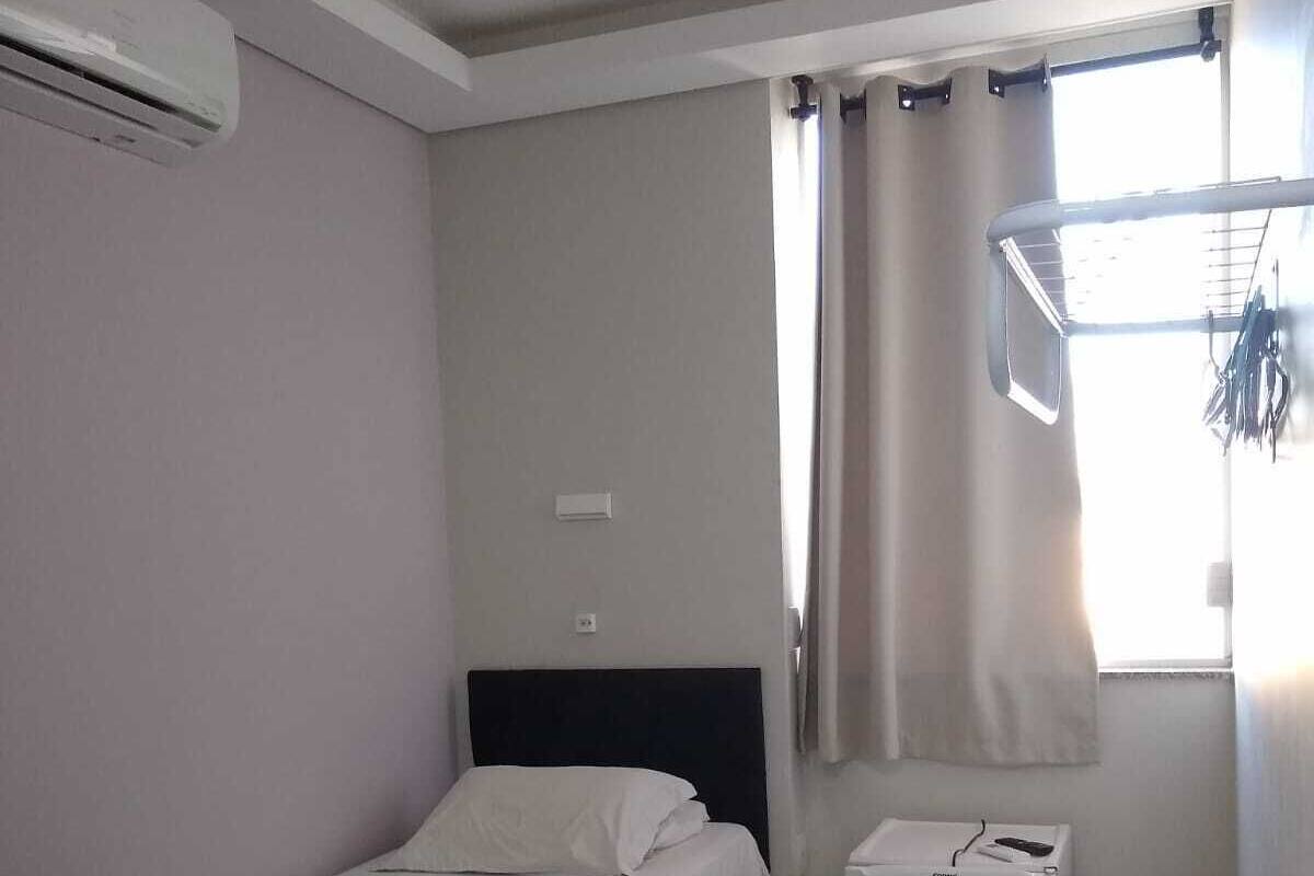 Quarto pequeno e limpo com cama, escrivaninha e cadeira. Há uma janela com cortinas, uma mesinha e um armário branco.