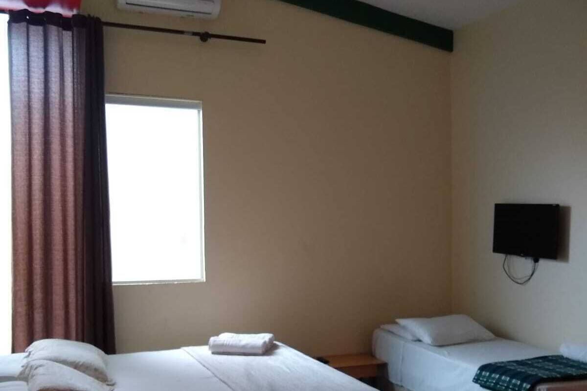 Quarto de hotel com duas camas, uma janela com cortinas, ar condicionado e uma televisão fixada na parede.