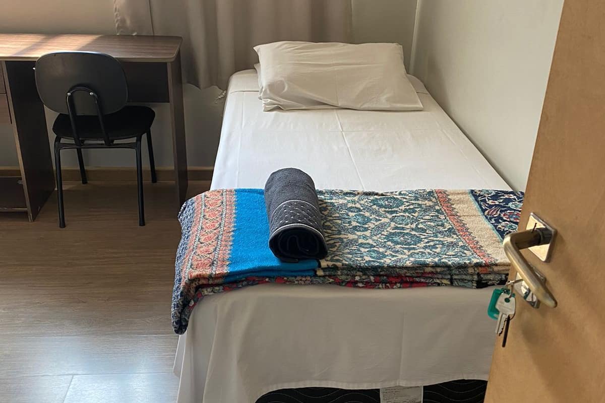 Uma cama de solteiro com um edredom estampado, uma escrivaninha com cadeira e uma janela com cortinas.