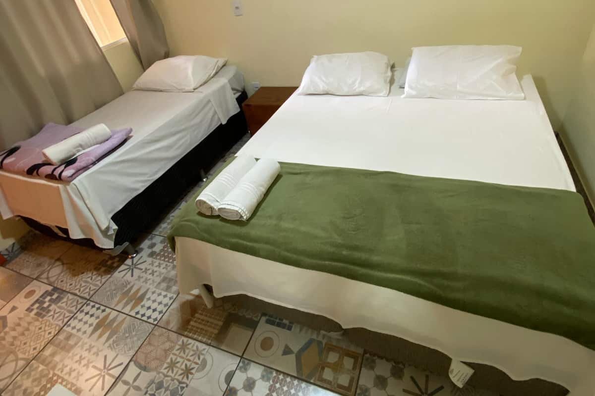 Quarto com duas camas, uma com manta verde e outra com roupa de cama branca. Há uma pequena mesa em primeiro plano e uma janela com cortinas.