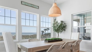 A dining area with a wooden table and wicker chairs, large windows with a view, and a hanging pendant light. - Oceanfront 6 Bedroom Heated Pool Home in Cinnamon Beach! Barefoot Sands!! (Palm Coast)