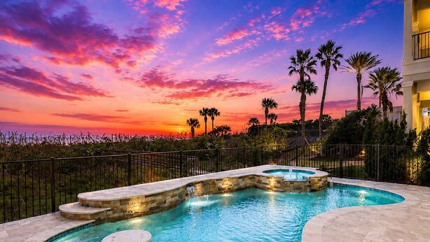 A swimming pool with a waterfall feature, surrounded by a fence and palm trees, with a sunset in the background. - Oceanfront 6 Bedroom Heated Pool Home in Cinnamon Beach! Barefoot Sands!! (Palm Coast)