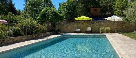 A rectangular swimming pool with clear blue water, surrounded by a stone wall and a tiled deck. There are two umbrellas providing shade, and a small building is visible in the background.