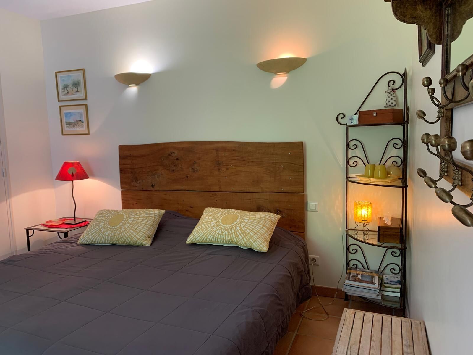 A bedroom with a wooden headboard, two bedside lamps, a red lamp on a nightstand, a shelf with decorative items, and a wall-mounted light fixture.