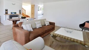 Ein modernes Wohnzimmer mit einem braunen Ledersofa, einem weißen Sessel und einem Glastisch. Im Hintergrund ist eine Küchenzeile mit einer Holztreppe zu sehen.