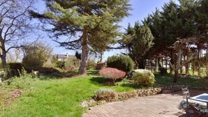 Ein gepflasterter Patio-Bereich mit Tisch und Stühlen, umgeben von einem Garten mit verschiedenen Sträuchern und Bäumen.